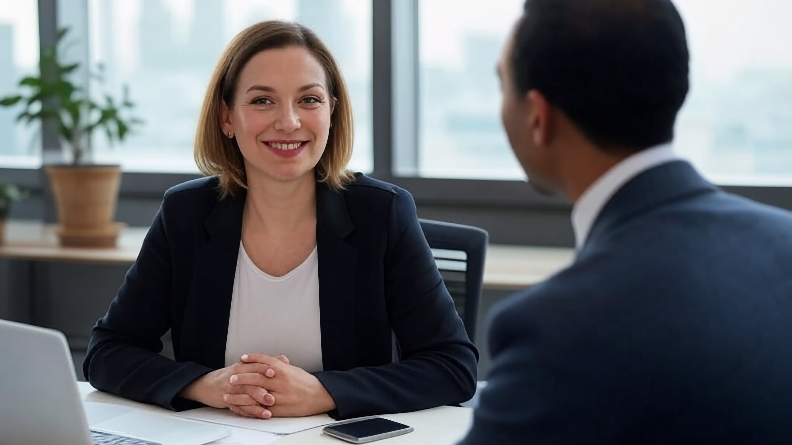 Femme souriante en blazer menant un entretien en face à face à travers un bureau avec un ordinateur portable