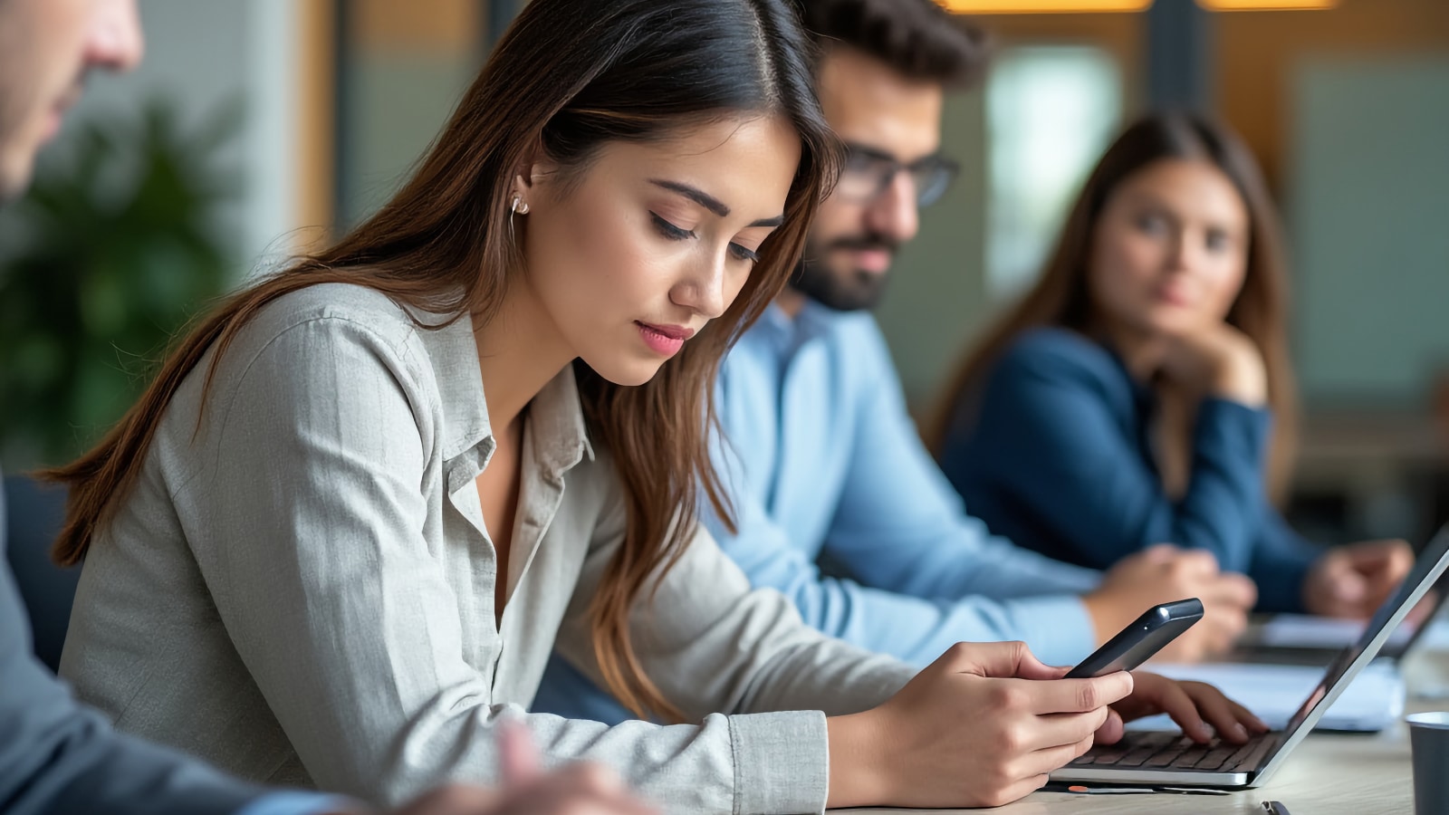 Jeune femme consultant son téléphone à une table de conférence avec des collègues et des ordinateurs portables en arrière-plan
