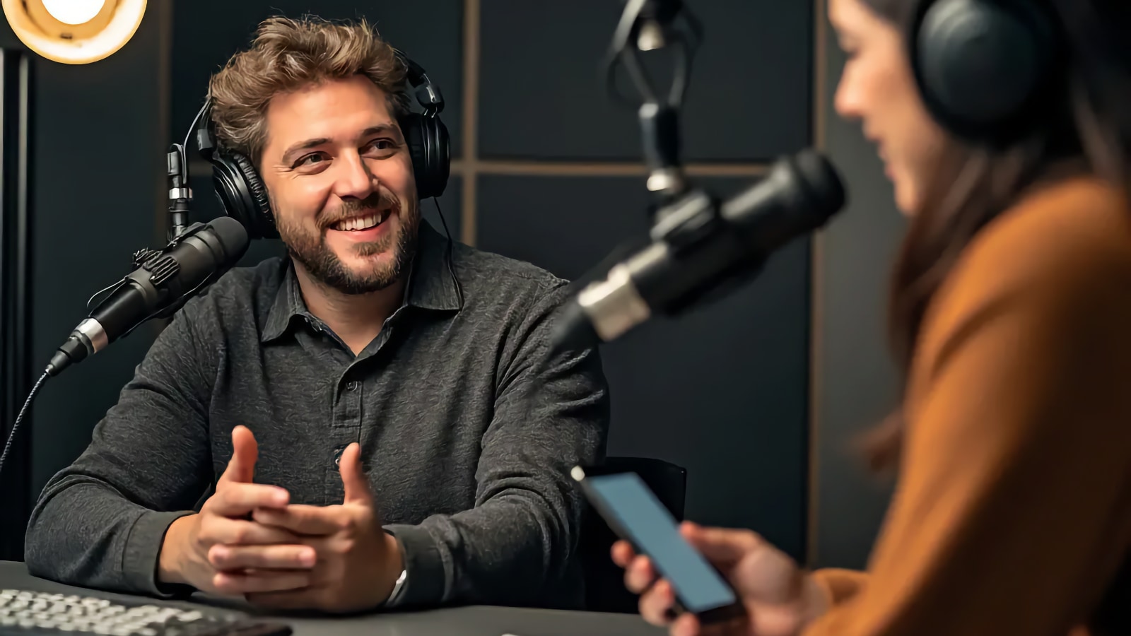 Homme portant un casque parlant dans un microphone de studio tandis qu'une femme tient un téléphone de l'autre côté du bureau
